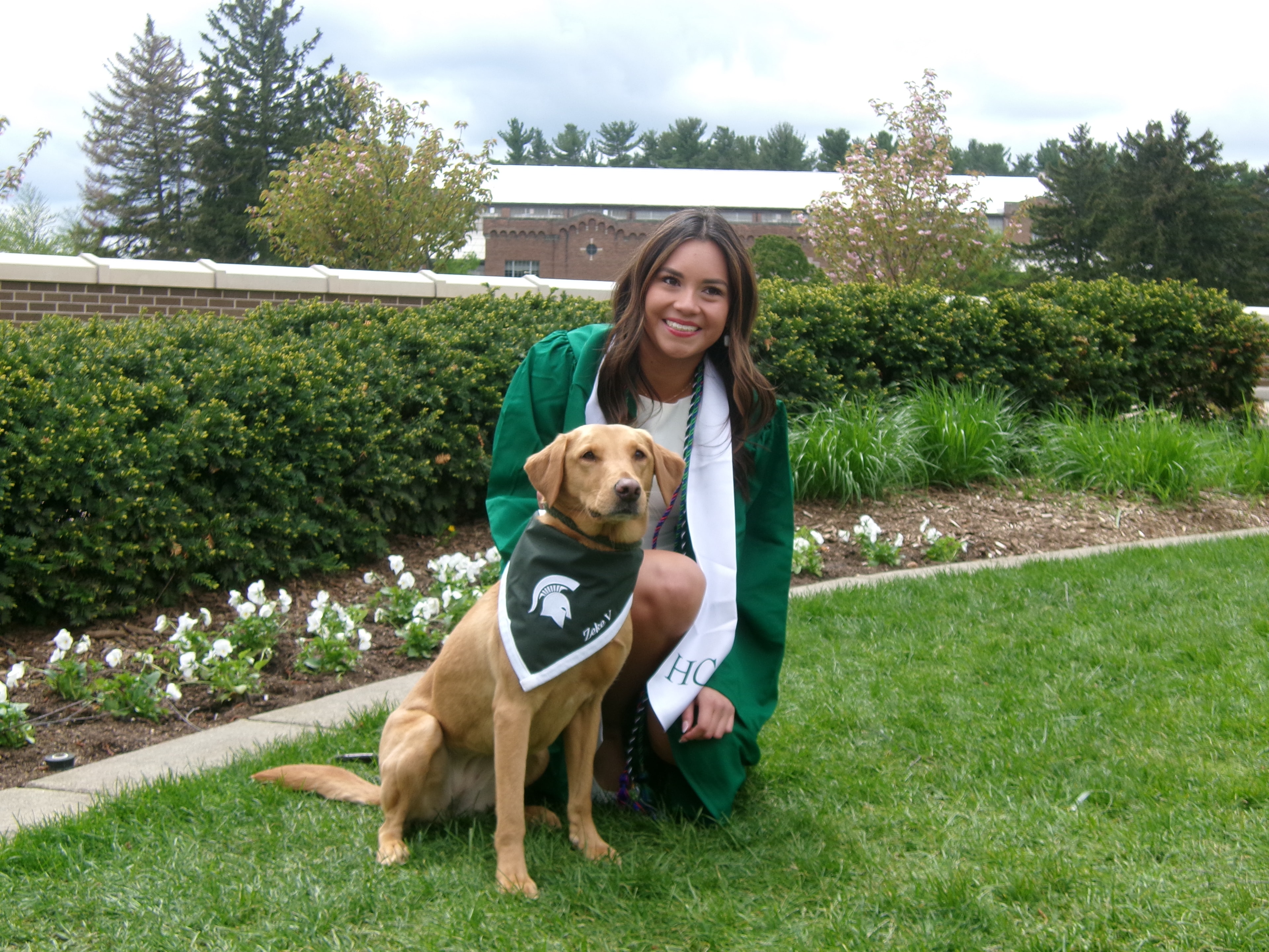 MSU graduate in green cap and gown kneeling with Zeke wearing his Spartan bandana